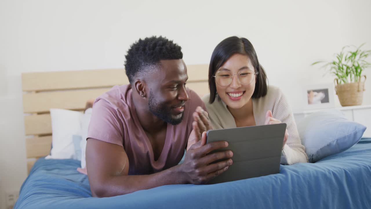 Happy diverse couple using tablet and lying in bedroom