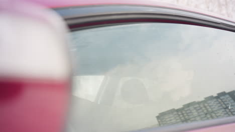 young student in sunglasses rolls up car window during snowy winter day as snowflakes drift through air and building reflects on windshield
