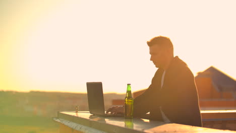 a male freelance programmer sits on a skyscraper roof with a laptop and beer typing code on a keyboard during sunset. remote work