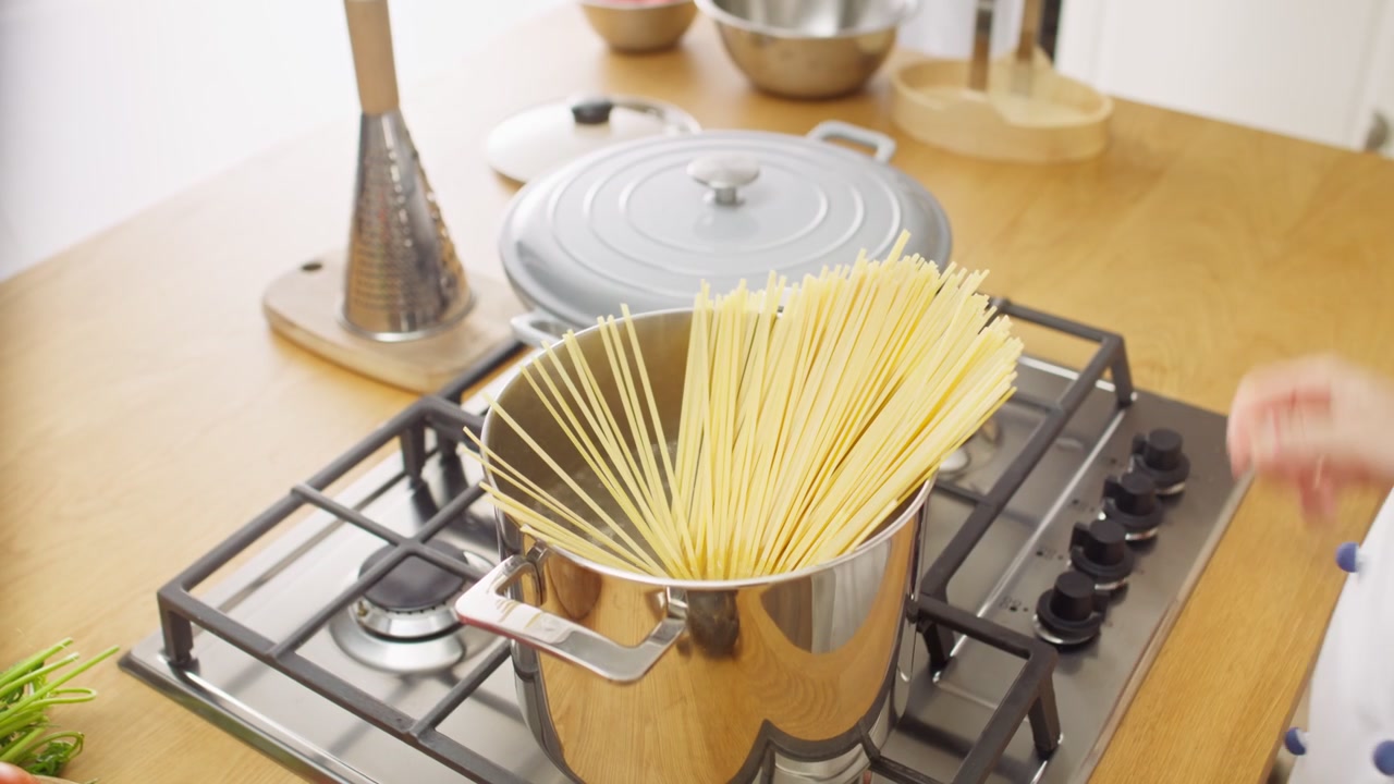 Mani che mettono spaghetti secchi in una pentola di metallo su un fornello a gas in una cucina luminosa