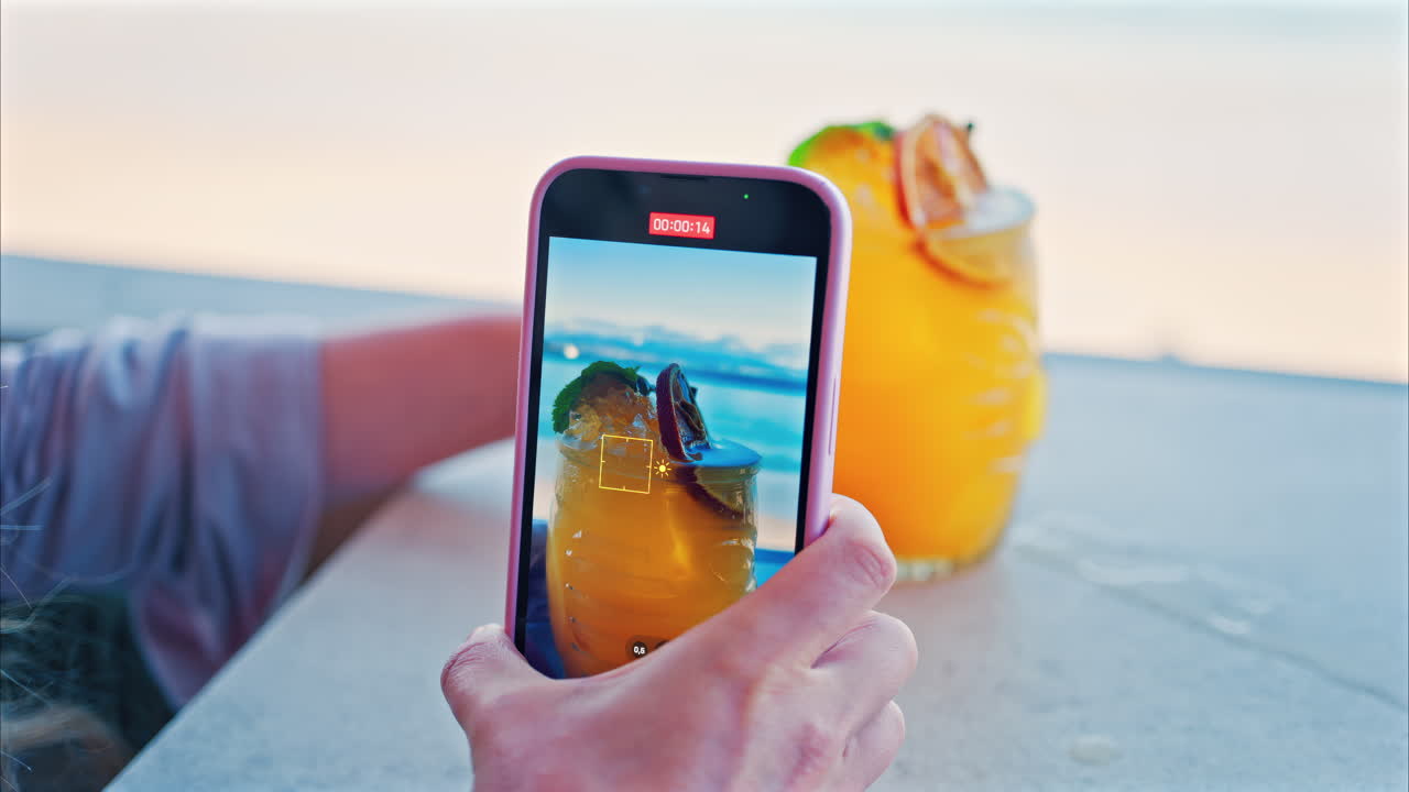 Close up of a woman's hand filming an orange cocktail on her phone with a blurred view of the sea on the background