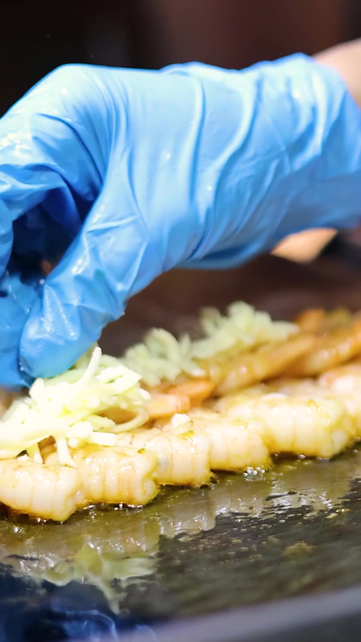 Una mano con guanto blu sparge formaggio grattugiato su spiedini di gamberi che grigliano su una piastra calda.