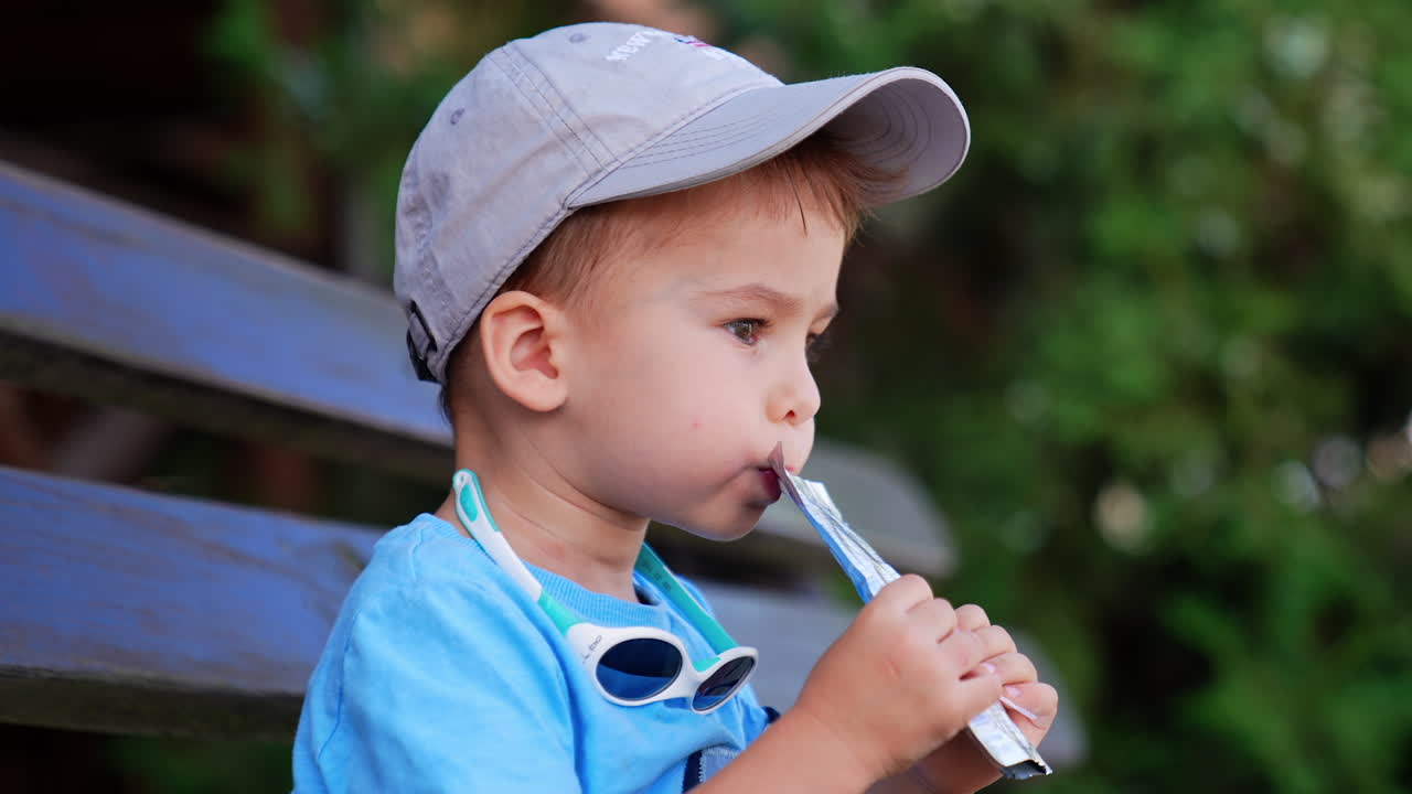 Mom's hand gives a doy pack to a baby boy in cap. Caucasian kid drinks juice from a straw. Close up. Blurred backdrop.