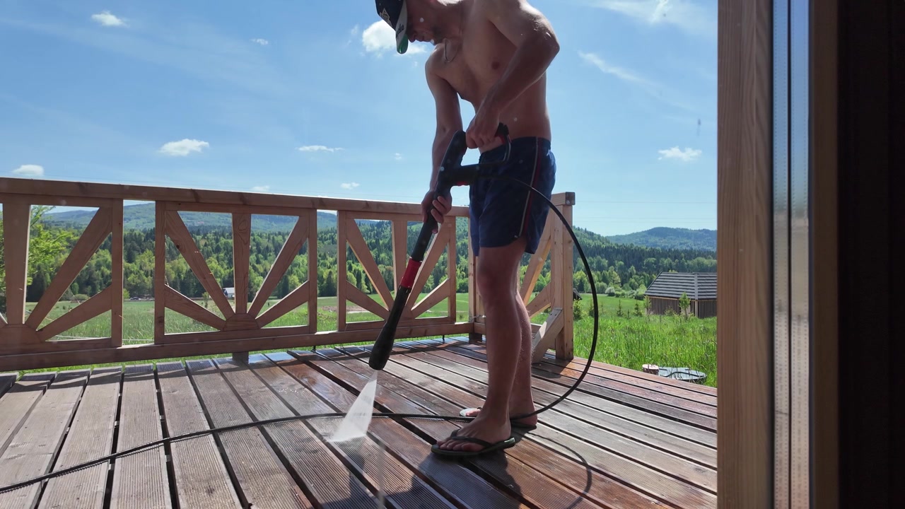 Hombre sin camiseta en pantalones cortos azules limpia a presión una terraza de madera en una cabaña de troncos en un día de verano soleado y brillante, rodeado de verdes prados, bosque y montañas.