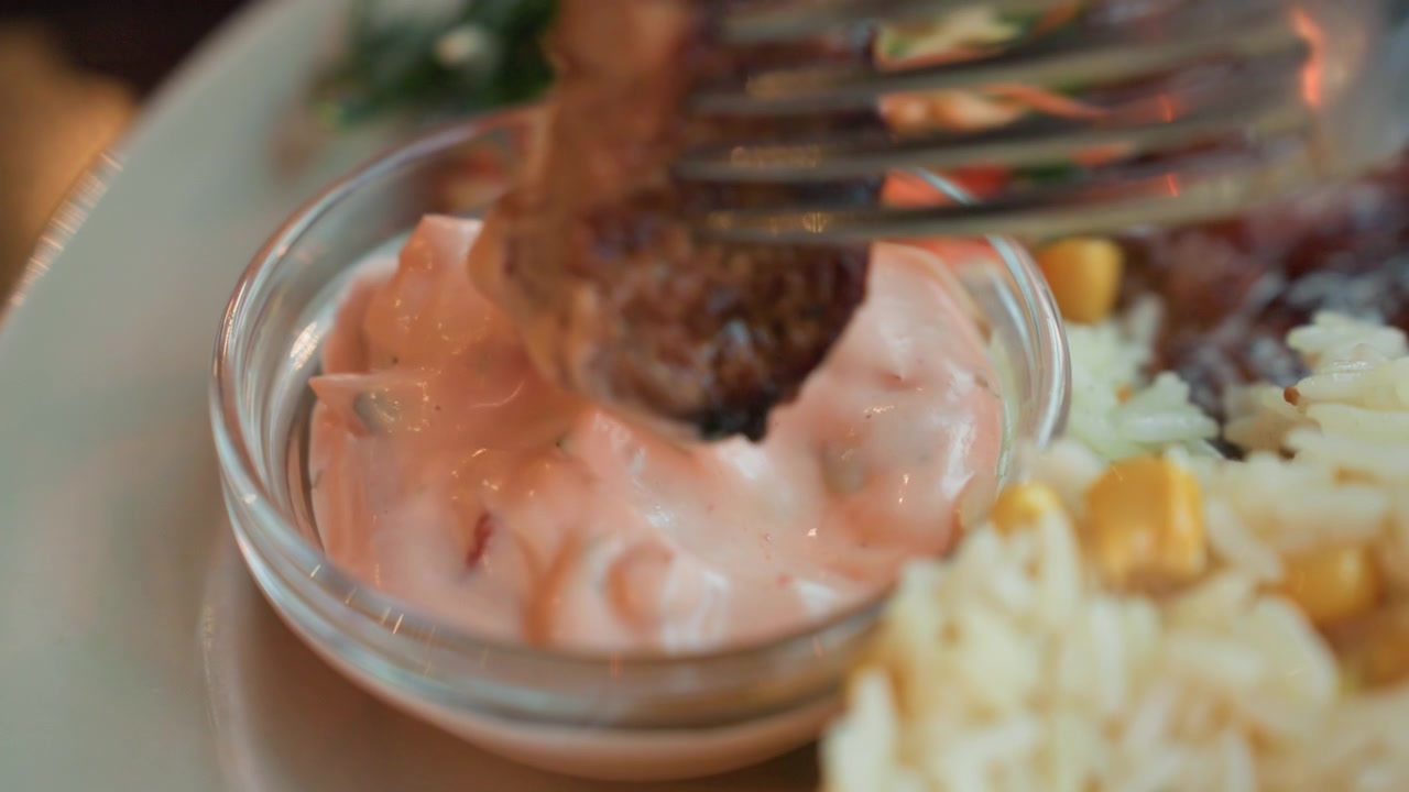 Close up view of pink creamy sauce in small glass bowl surrounded by rice and vegetables. fork dips meat into thick textured condiment throughout sequence
