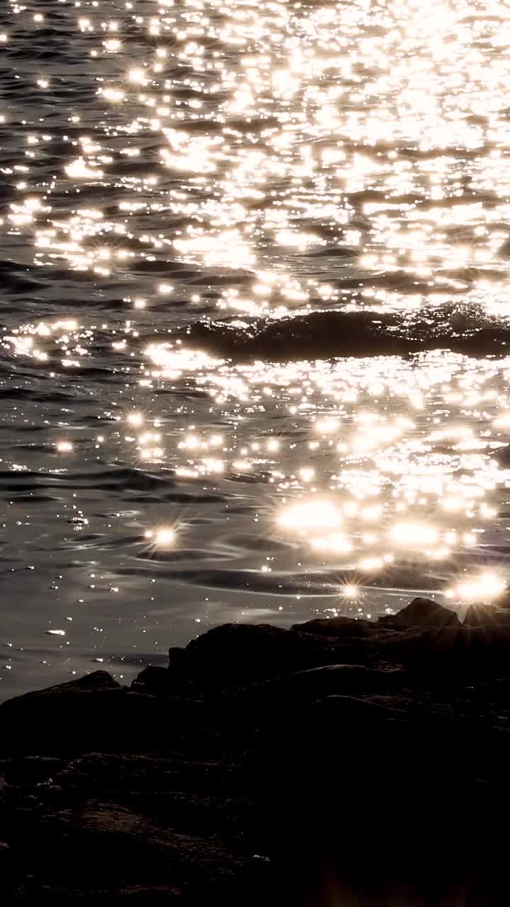 Bright sunlight creates a shimmering bokeh effect on gentle sea waves hitting a rocky shoreline