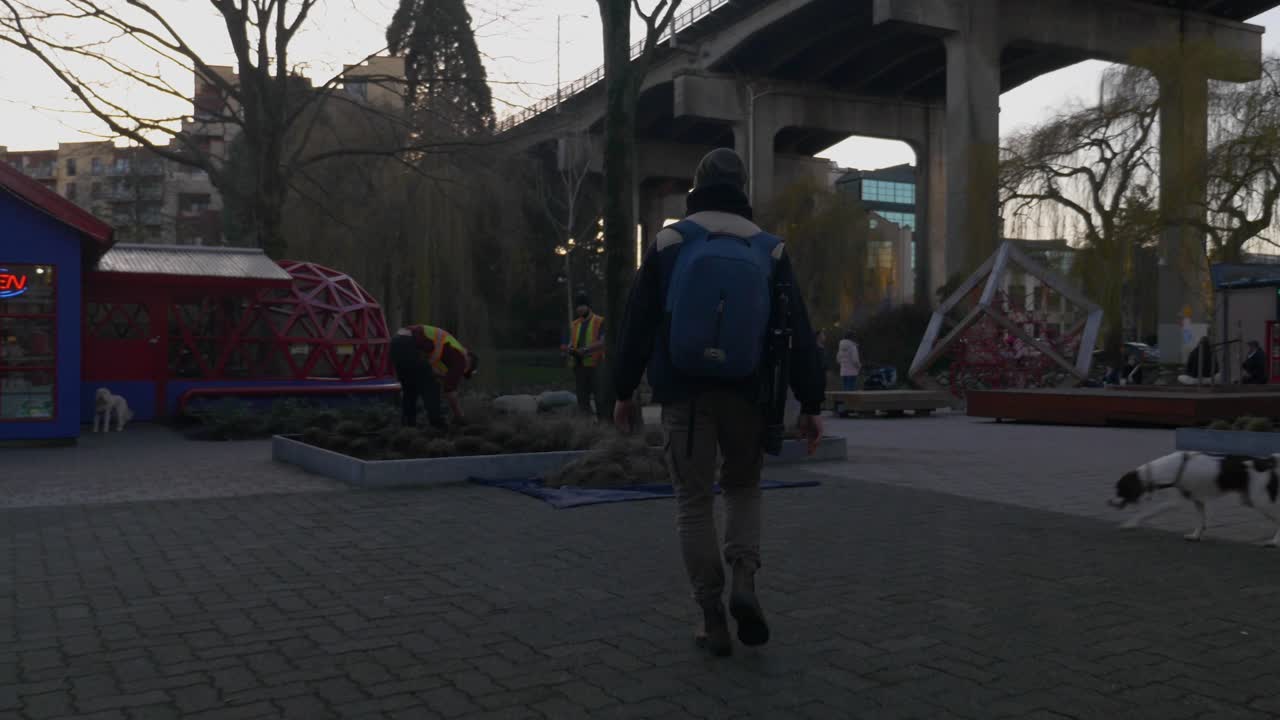 Man with a backpack walking on a pavement in an industrial site passing by a dog and people working under the city bridge