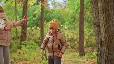 young girl joyfully tosses dry autumn leaves into air as smiling mother joins her, both dressed warmly in colorful forest, surrounded by falling foliage and vibrant greenery