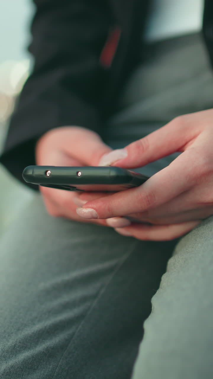 Close up of woman operating phone with both hands while seated in formal attire, wearing white wristwatch, with soft bokeh background of cityscape and people walking