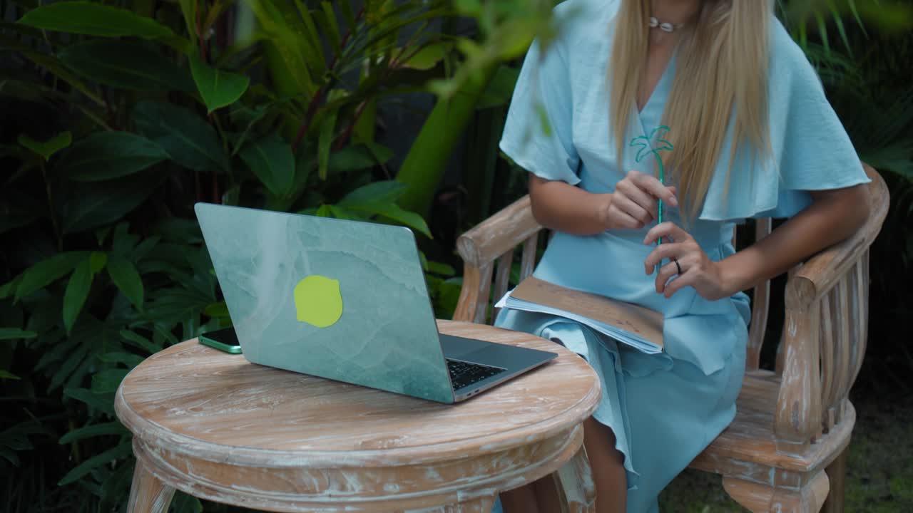 una chica en un vestido azul remotamente en línea trabajando detrás de una computadora portátil y mirando a la pantalla el patio trasero con plantas verdes