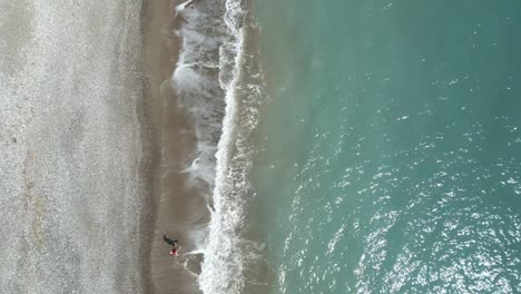 A-single-person-walking-on-pissouri-beach-in-cyprus,-waves-lapping-at-the-shore,-aerial-view