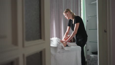 massage therapist in black attire performing chest massage on relaxed female client lying under white sheet in bright spa room with grey curtains in background, captured from side doorway angle