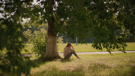 woman sits on grassy ground under shaded tree, thoughtfully resting hand on her leg, surrounded by greenery, background includes park setting with poles and open field