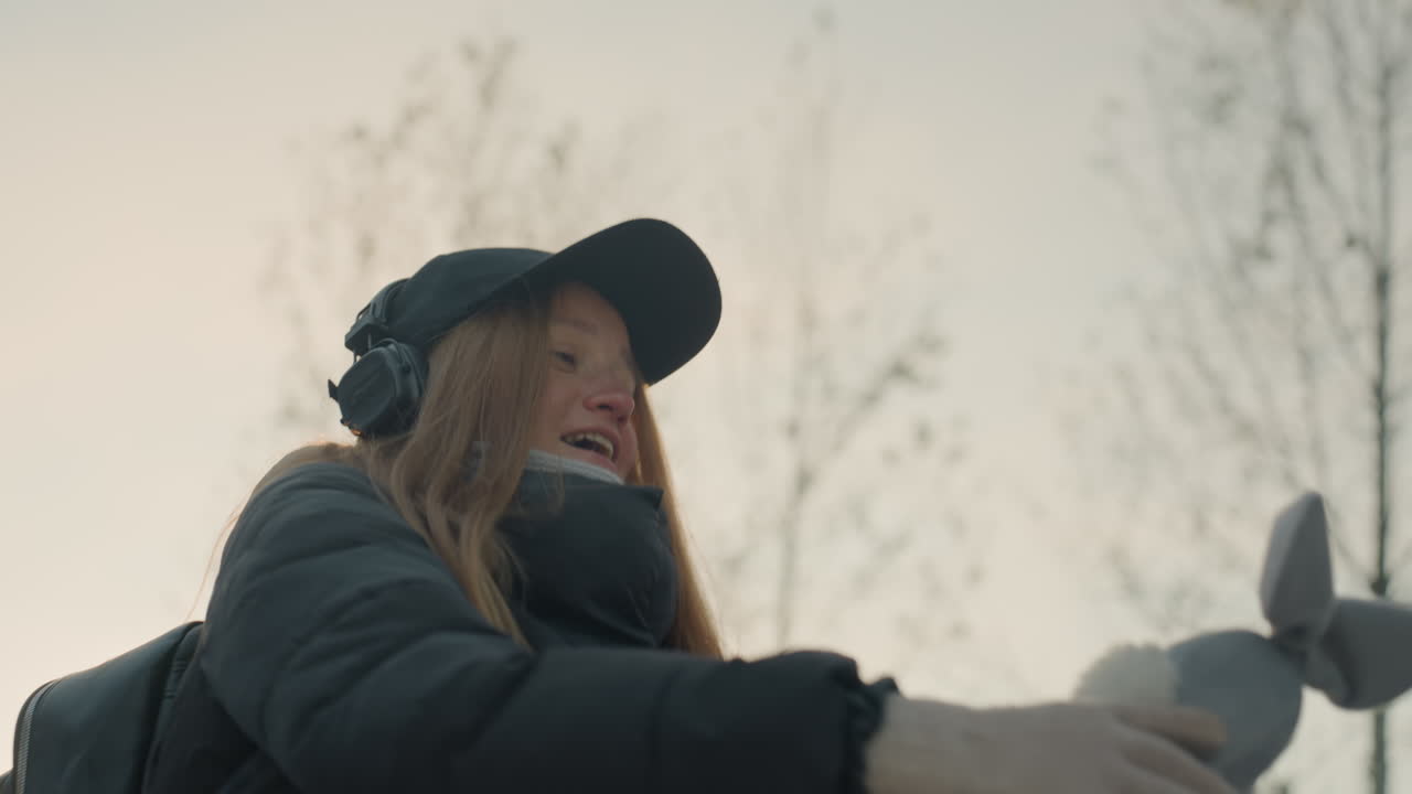 Donna caucasica dai capelli rossi che tiene un coniglio di peluche vicino al tramonto in un parco urbano, cappotto invernale, berretto e cuffie, zaino a tracolla, mani guantate che accarezzano il giocattolo di peluche, sorriso gioioso e risata sommessa
