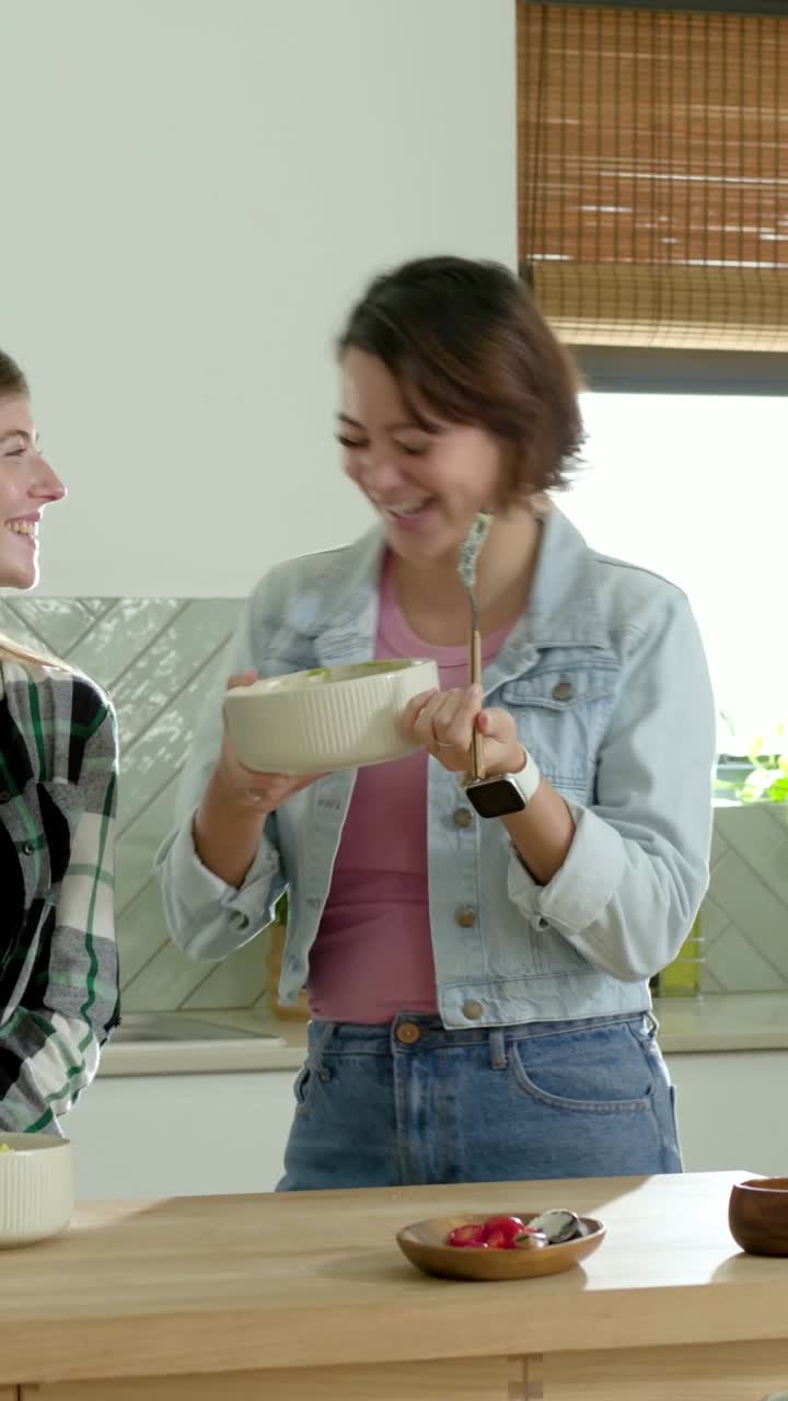 Video verticale: Donna in denim che assaggia un cucchiaio all'isola della cucina dopo che l'amica ha mescolato la ciotola.