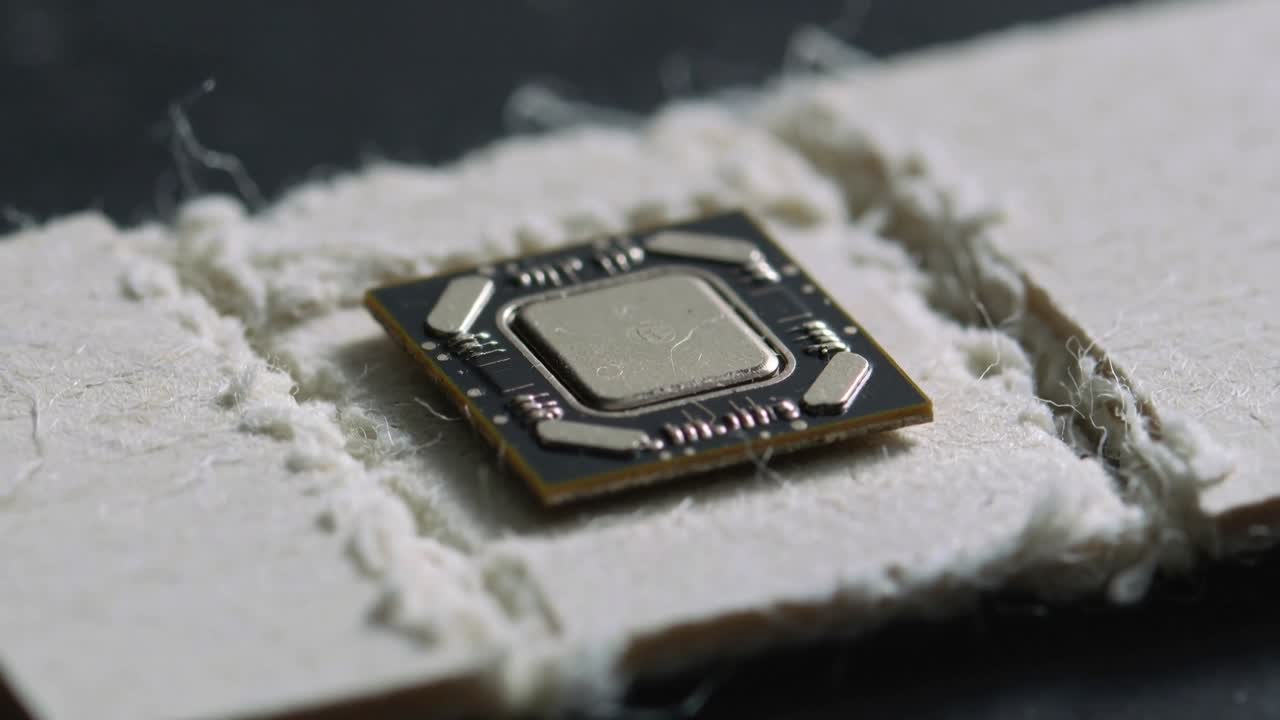 Fotocamera macro che ingrandisce, rivelando un chip quadrato con cappuccio metallico e tracce di saldatura su una striscia fibrosa strappata.
