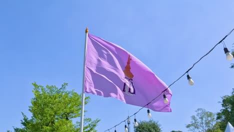 a purple flag waves gently against a clear blue sky, surrounded by green foliage and decorative string lights.
