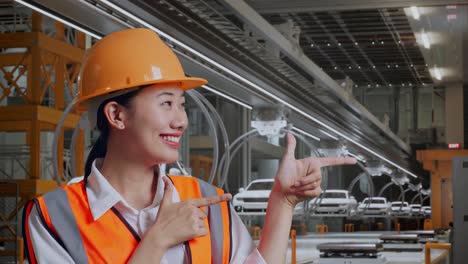 close-up de ingeniera asiática con casco de seguridad sonriendo y apuntando a un lado mientras está de pie en la fábrica de automóviles ev