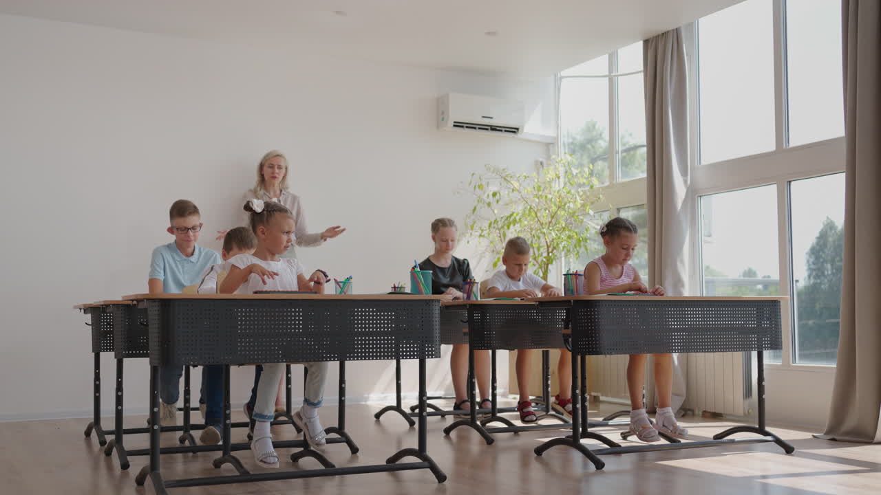 the children in the class are sitting at their desks and the teacher walks around the class and explains the topic of the lesson. Smart Children Learning in Friendly Modern Environment