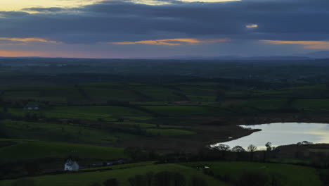 Time-lapse-of-rural-farming-landscape-of-farmhouse-at-lake-in-grass-fields-and-hills-during-dramatic-cloudy-sunset-viewed-from-Keash-caves-in-county-Sligo-in-Ireland