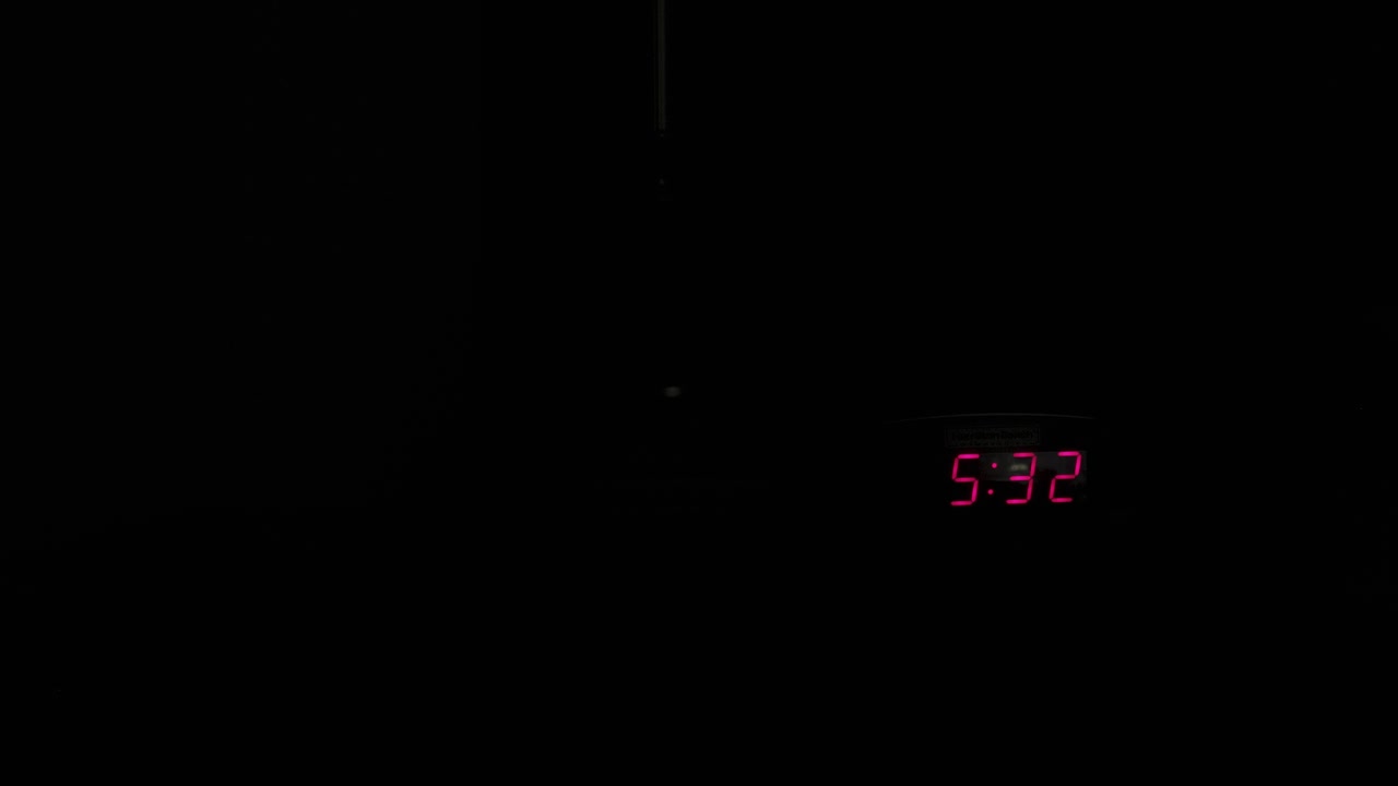 In a dark room, an alarm clock glows on a nightstand. A person turns on the light and shuts off the alarm, ideal for morning, sleep, and lifestyle visuals