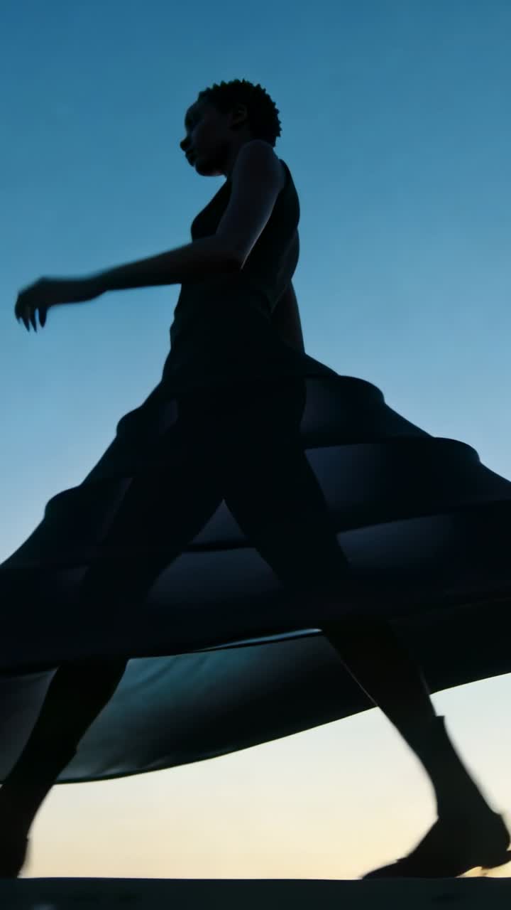 Vertical video: Walking woman after low-angle framing moving across shoreline at dusk, flared skirt