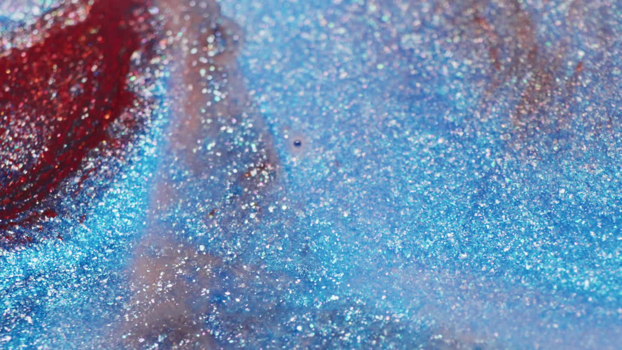 A macro shot captures red and blue glitter as water is injected and flows through, displacing the particles in shimmering motion