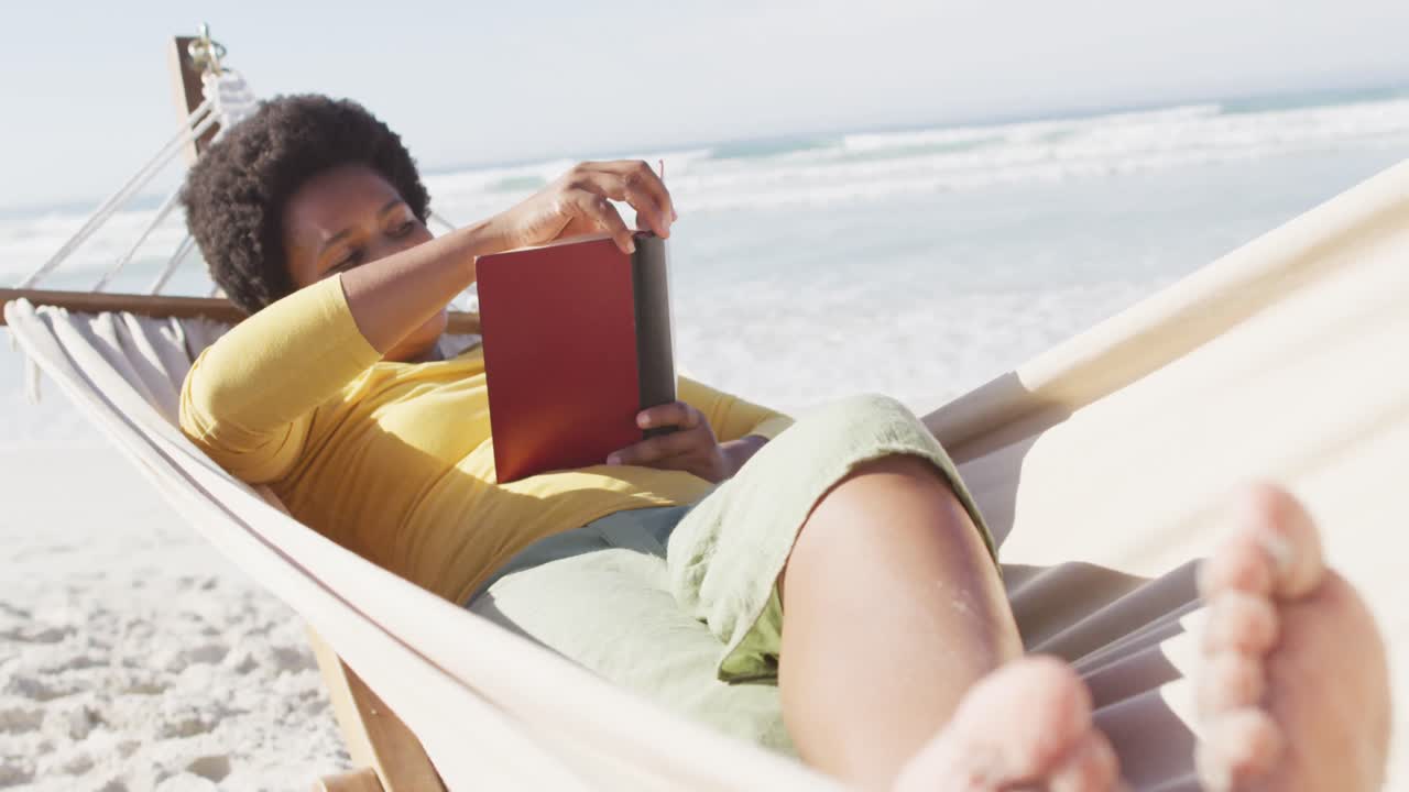 feliz mujer afroamericana leyendo y acostada en una hamaca en una playa soleada
