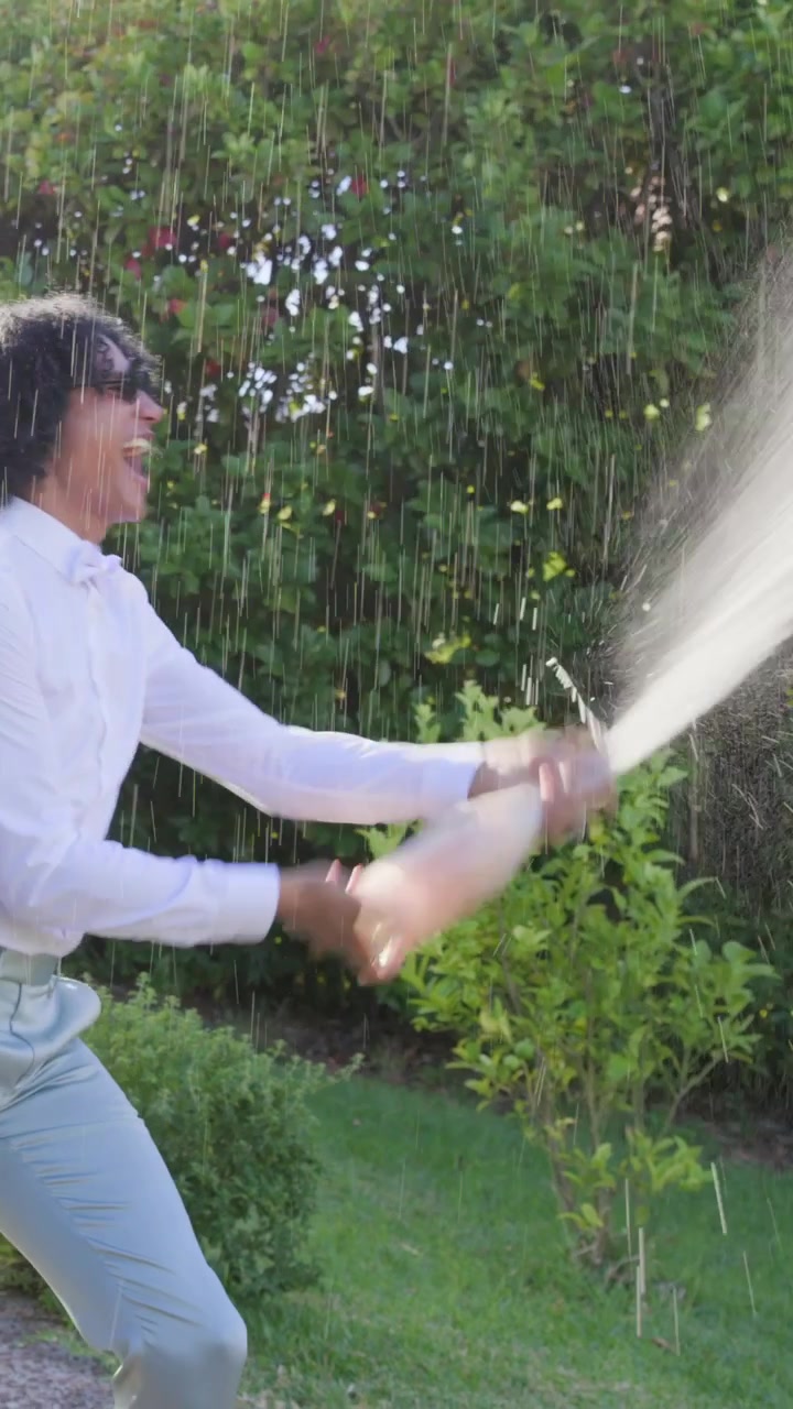 Vídeo vertical: Casal caminhando celebrando chegada ao jardim, estourando garrafa, borrifando, com roupas leves.