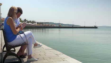 Man-and-woman-using-pad-sitting-on-waterfront-bench