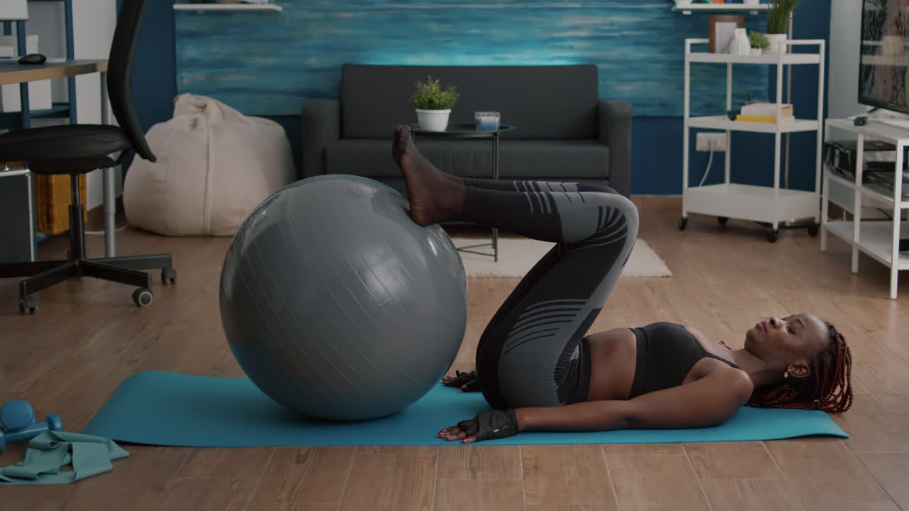 Donna di colore atletica che allena gli addominali sulla fitball nel soggiorno.