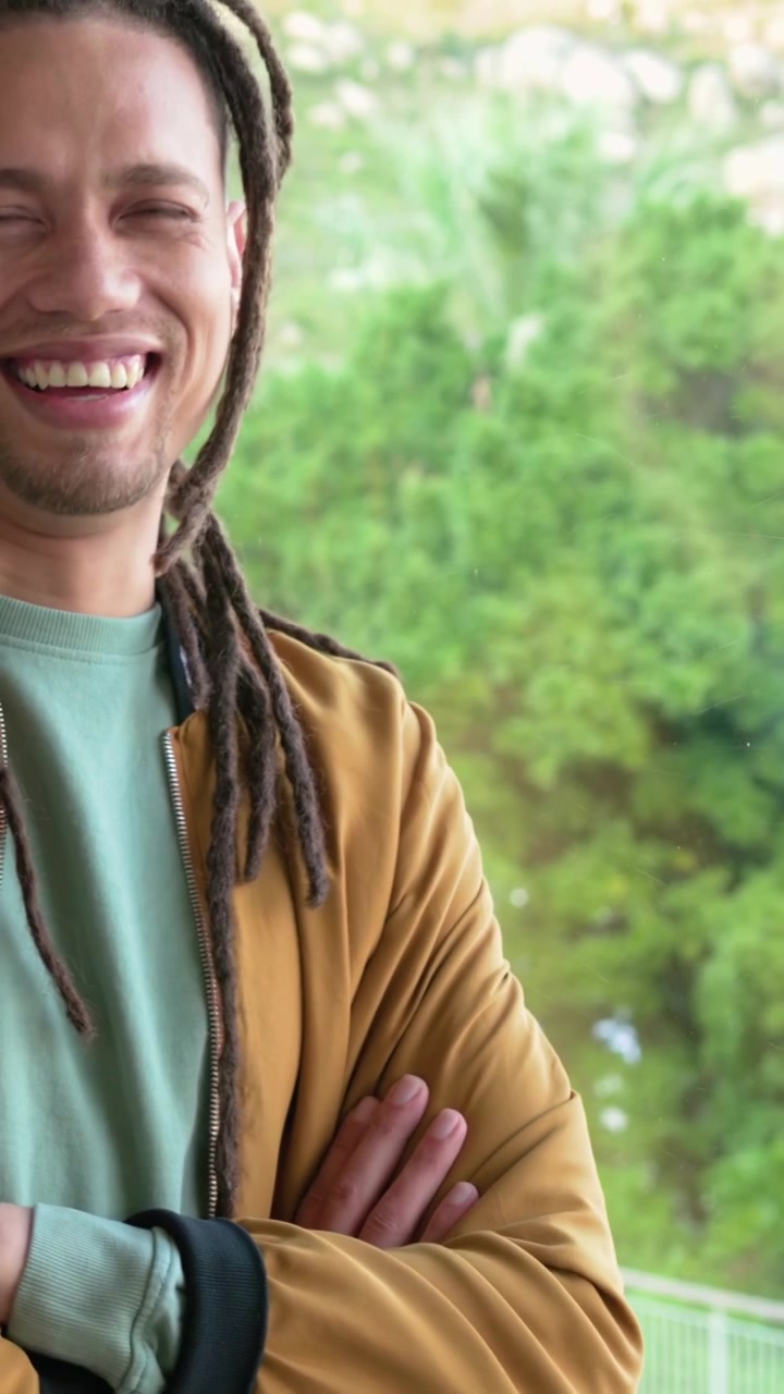 Vertikales Video: Kamera zoomt heran, Mann wechselt den Blick und lacht nahe einer Glastür in senfgelber Jacke.