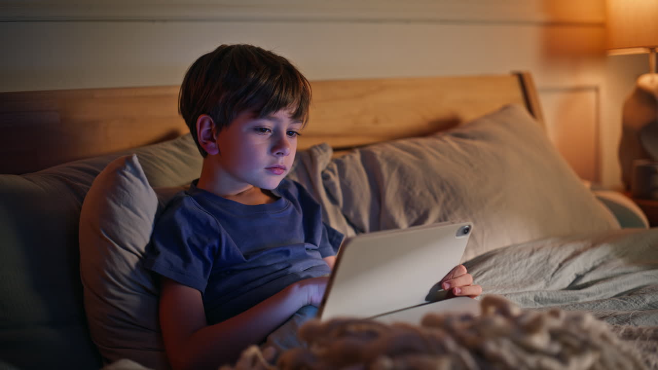Boy browsing tablet night bedroom closeup. Little kid focused computer lying bed