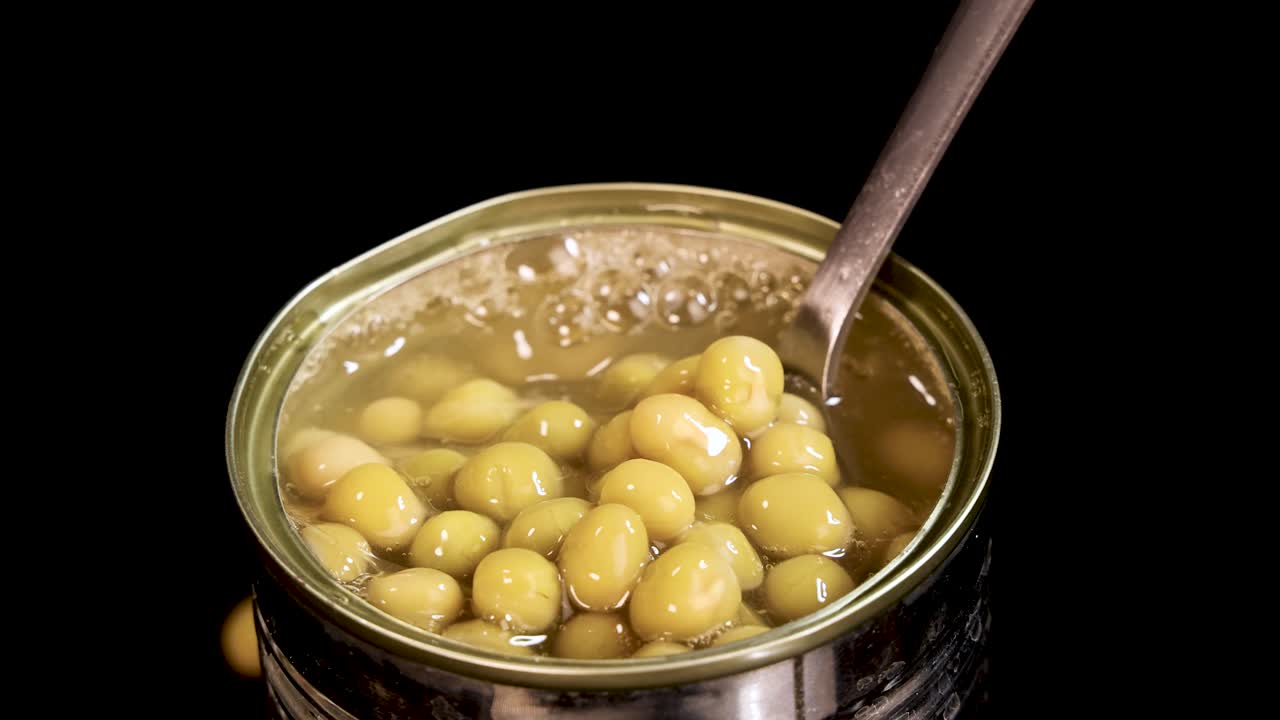 A metal spoon lifts and lowers cooked green peas in brine from an open can, under bright studio lighting with a black background