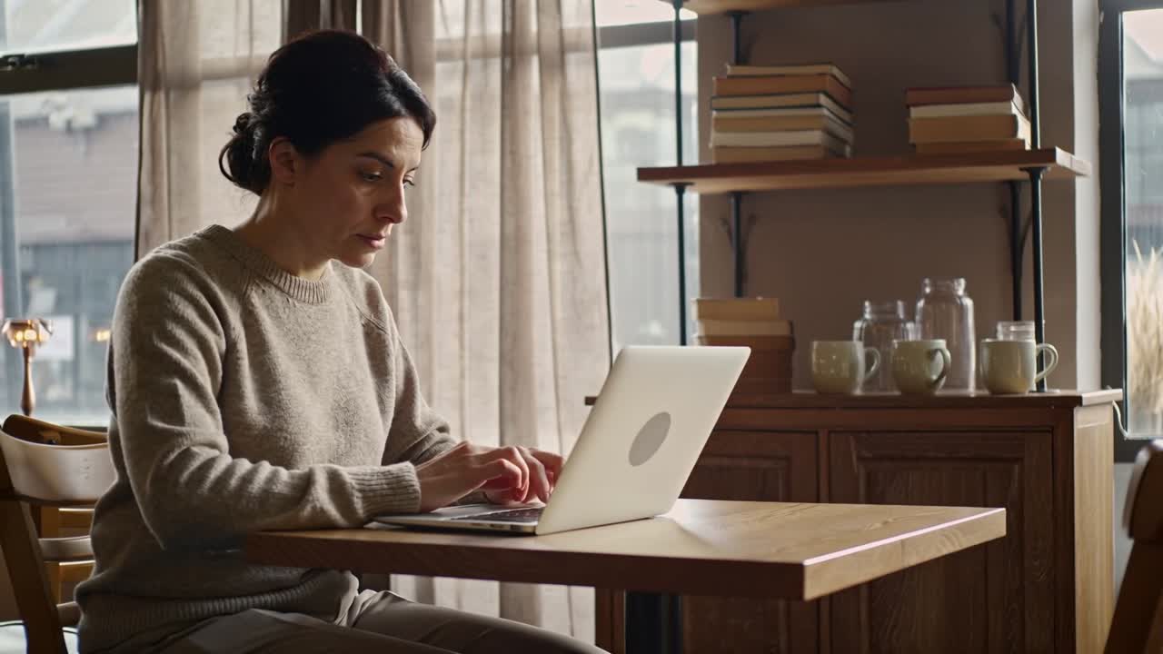 Entering frame from right, woman in sweater opening laptop and typing draft at home office nook