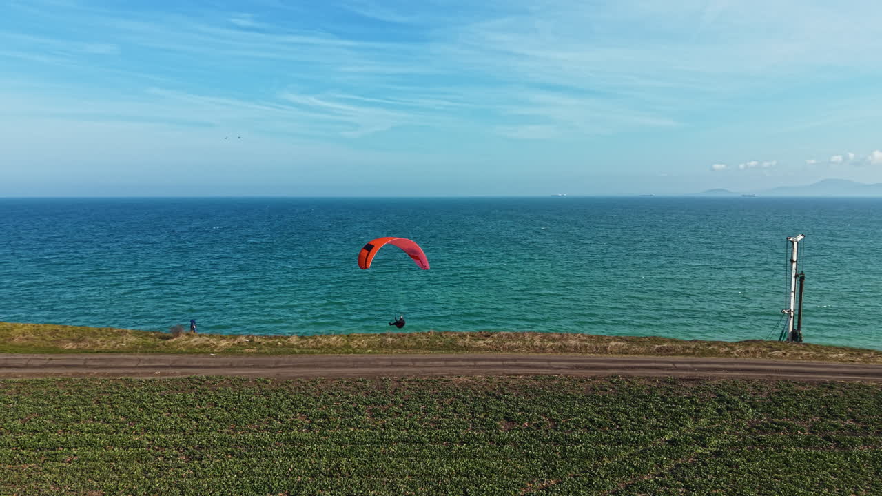 High above the coast, a person glides through the sky with a bright orange kite near a blue ocean on a sunny day