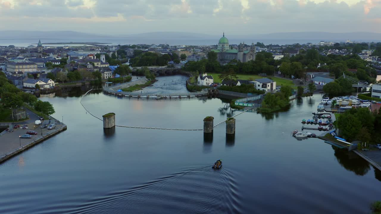 Aerial orbit around small boat near Salmon Weir with Galway cathedral behind at sunset