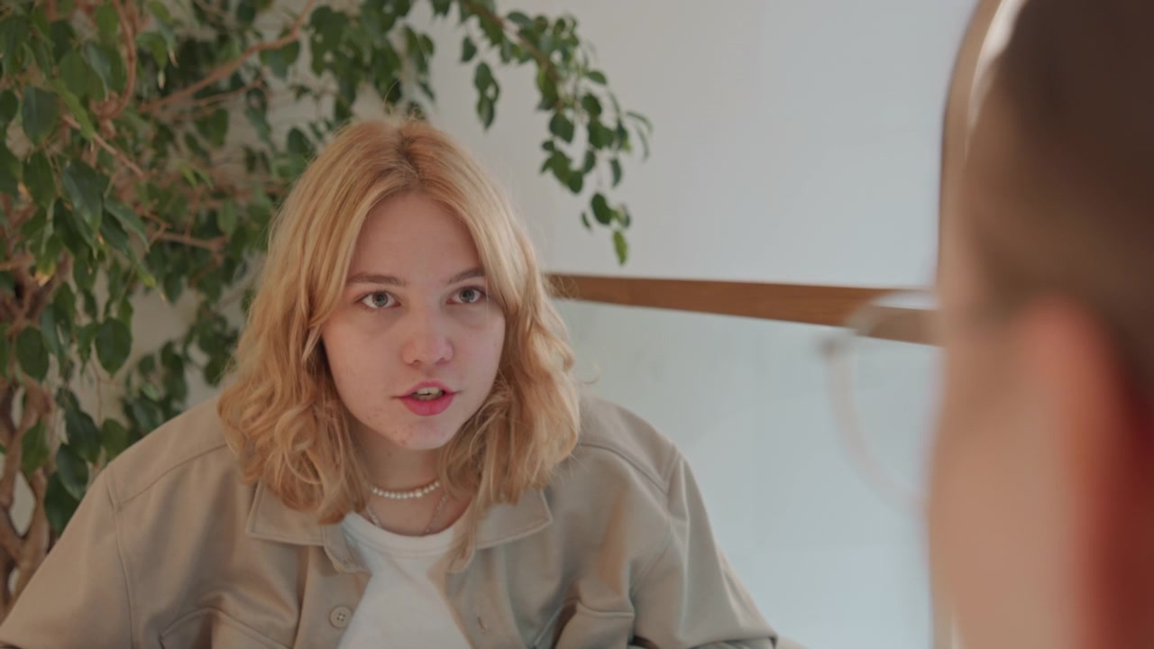 Close up of young woman with wavy blonde hair wearing beige jacket and white shirt engaged in conversation indoors. Woman is seated near green plants under soft lighting that creates warm