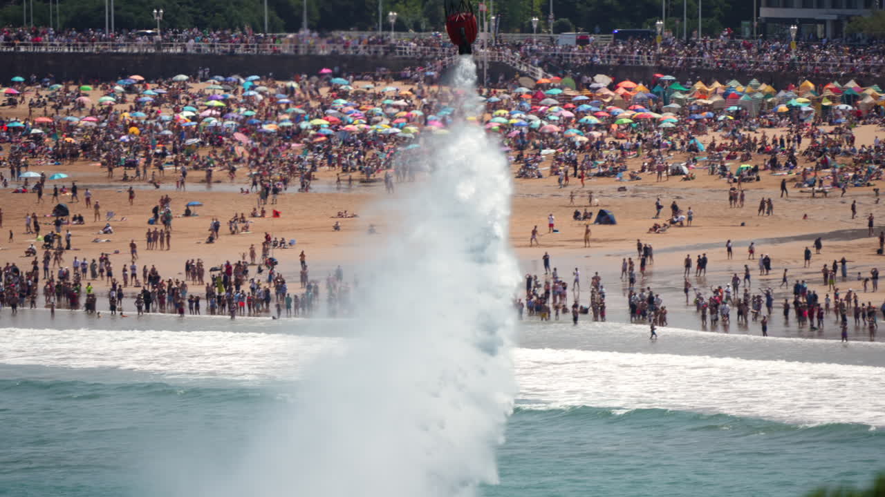 Yellow firefighting helicopter with bambi bucket drops water near crowded beach during public event with large audience and umbrellas
