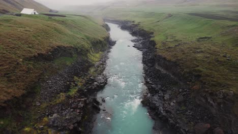 drone shot of a raging icelandic river and green farm fields above the black rocks and rapids