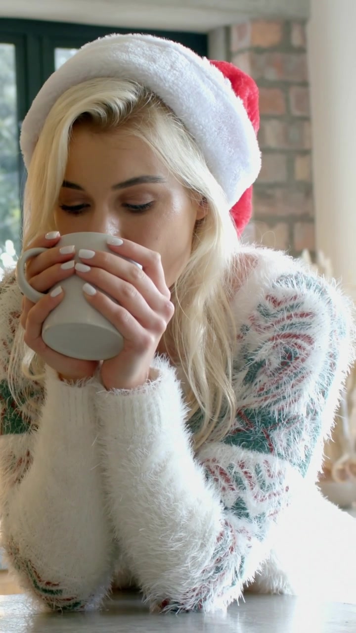 Video verticale: Donna che tiene una tazza, inala il vapore e sorseggia al bancone, indossando un cappello da Babbo Natale e un maglione
