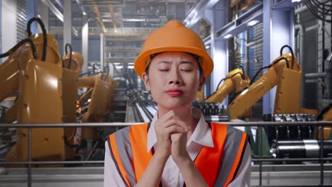 close up of asian female engineer with safety helmet pray for something while standing with industrial robotic arms working