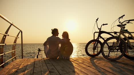 A-happy-couple,-a-guy-in-a-light-shirt-and-a-girl-in-a-straw-hat,-look-at-each-other-and-sit-on-a-beach-covered-with-boards-near-the-sea-at-sunrise.-A-guy-and-a-girl-came-to-the-beach-on-bicycles