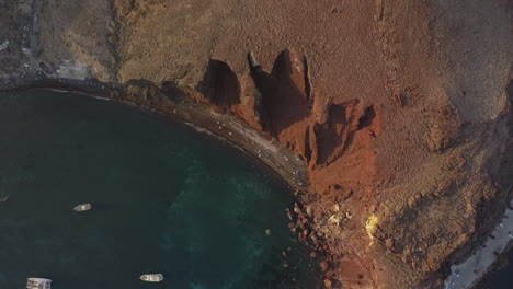 Deep-shadows-outline-eroded-red-cliffs-of-Santorini,-afternoon-aerial