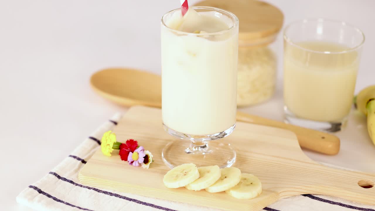 Hand stirs banana milkshake in glass, bright kitchen lighting, close-up, fresh fruit garnish