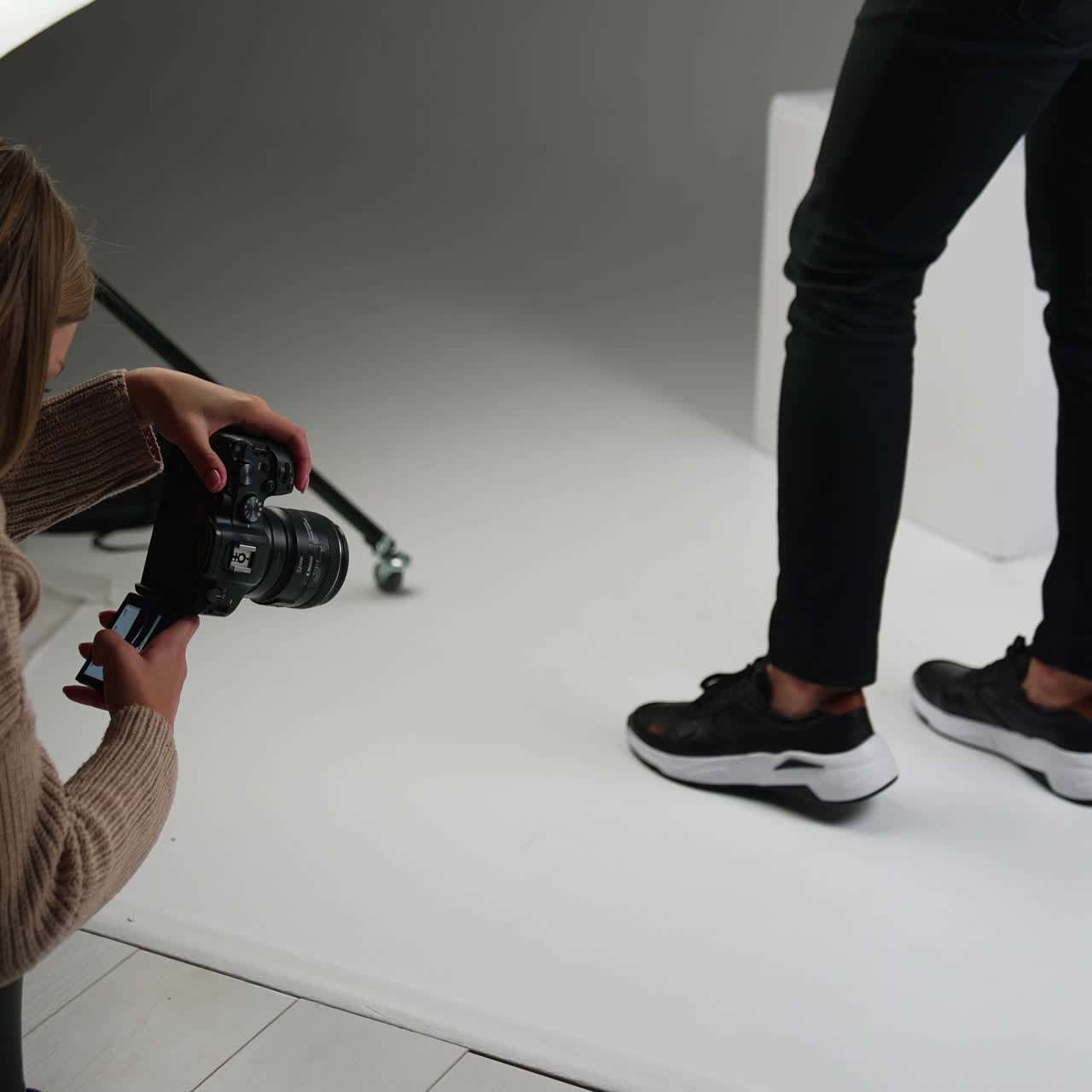Lady taking shot of male feet in new modern black sneakers with a white sole. Backstage work of a photographer in studio