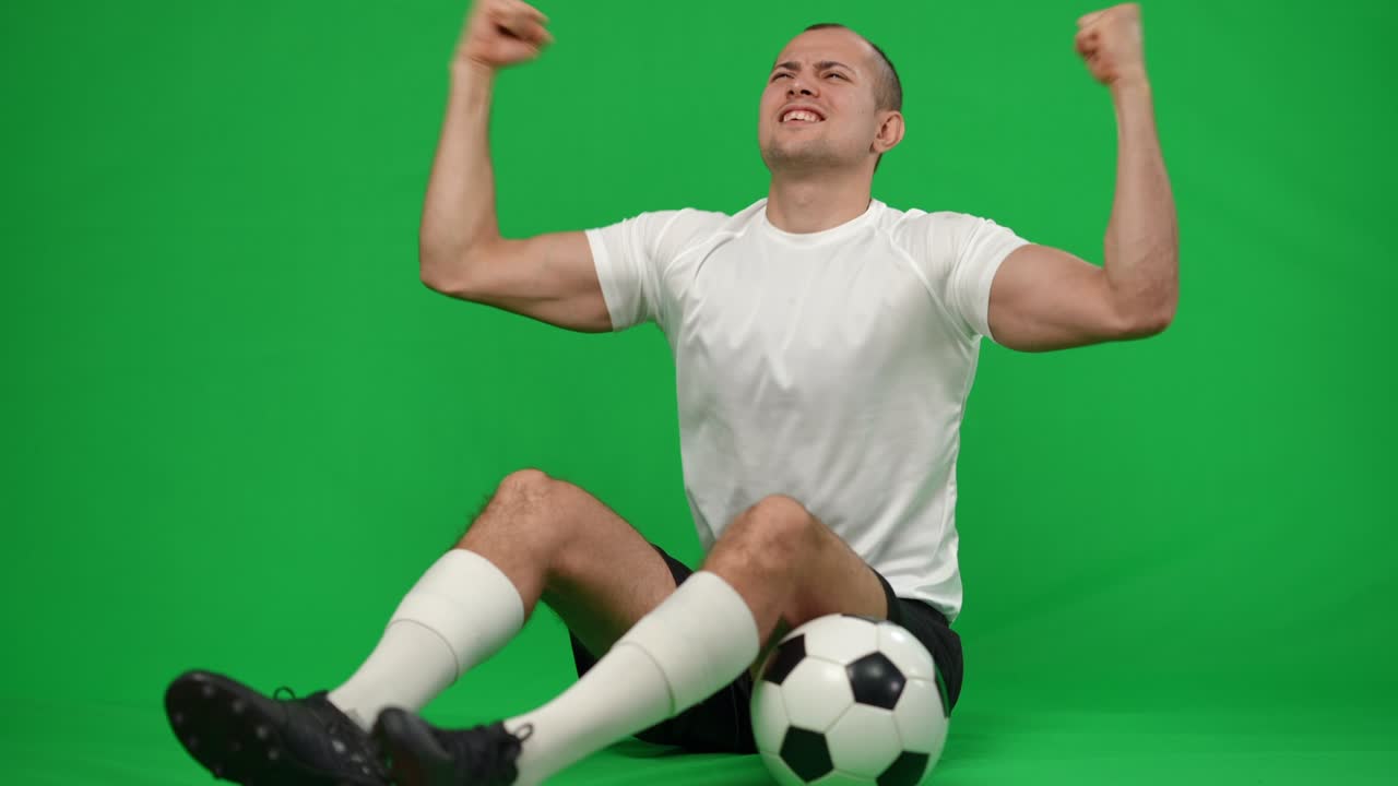 만족하고 흥분한 백인 스포츠맨이 크로마케이 배경에 축구공을 들고 앉아있으며, 녹색 스크린에서 성공을 기하는 행복하고 미소 짓는 젊은 축구 선수의 초상화입니다.