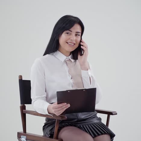 portrait of dark-haired woman talking the phone. beautiful young business woman sitting in the chair and holding black folder in the studio.