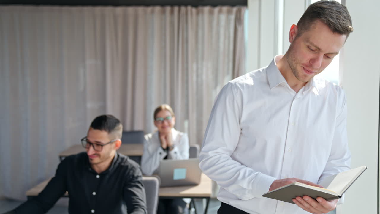 Handsome fit businessman looking at paper notebook stands at window. Happy smiling office staff sitting at desks at backdrop in blur.