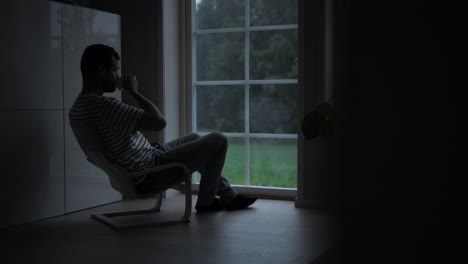 man in dark room watching rain through window at overcast day